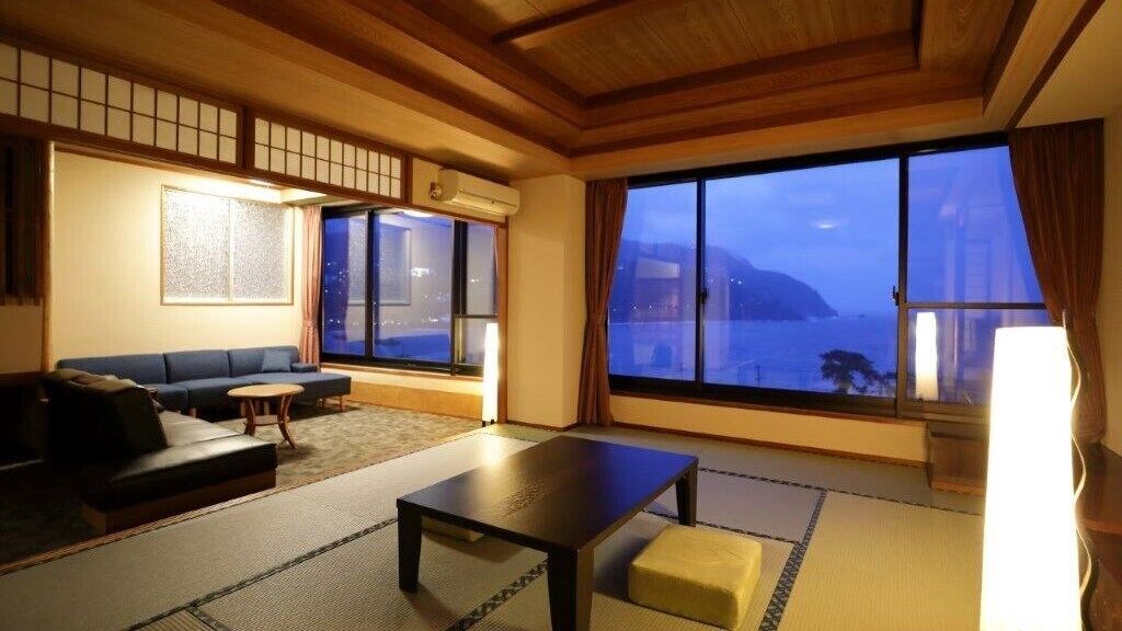 A traditional Japanese room with tatami flooring, a sofa, a table, and a large window with a view of the sea.