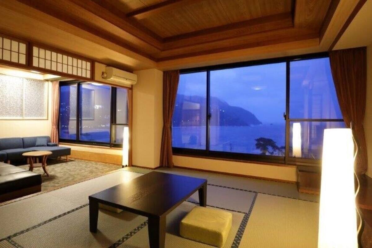 A traditional Japanese room with tatami flooring, a sofa, a table, and a large window with a view of the sea.