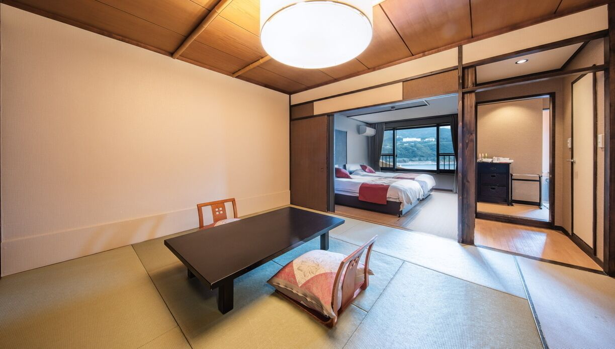 A traditional Japanese-style room with tatami flooring, a low table, and a sliding door leading to a bedroom with a bed and a view.