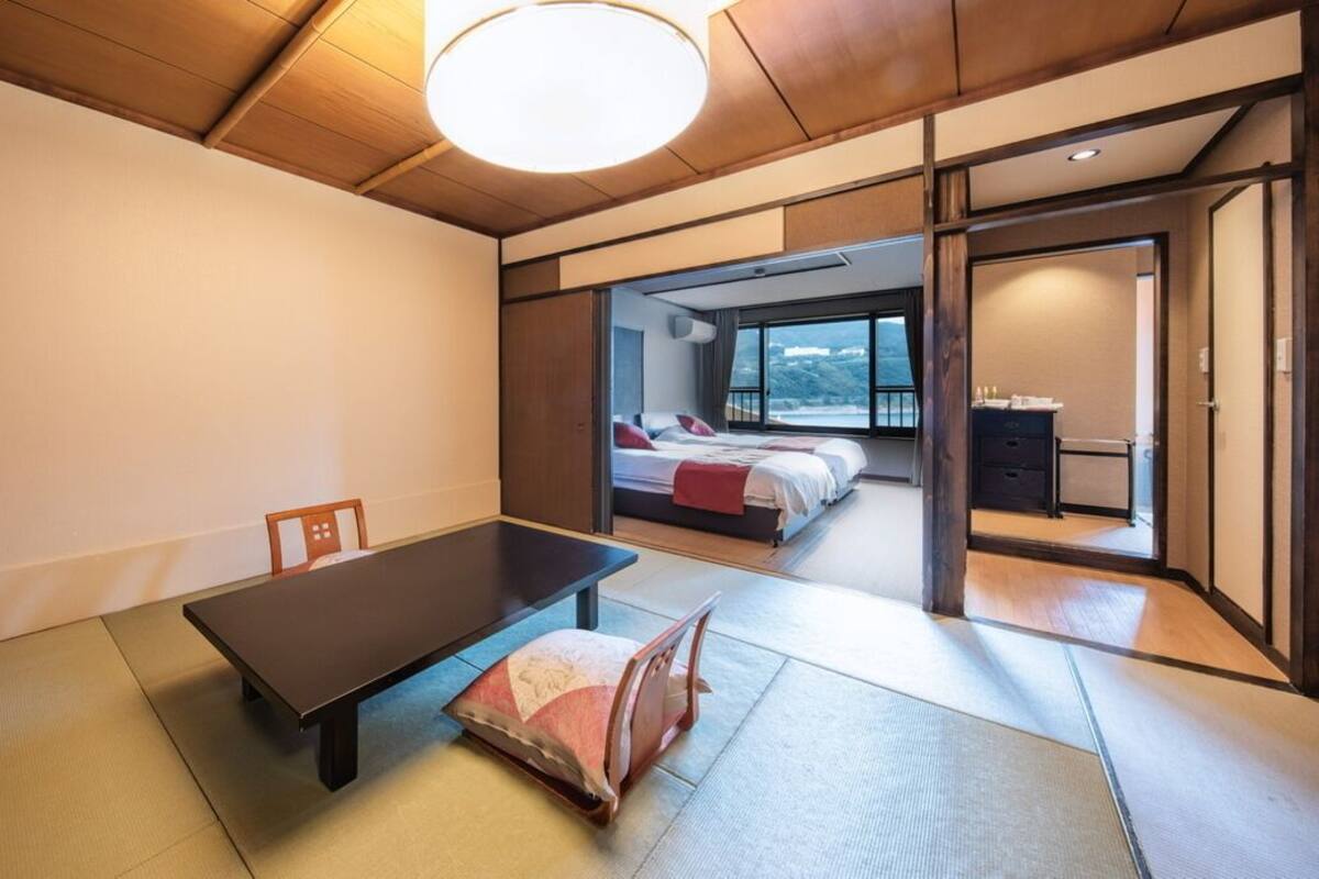 A traditional Japanese-style room with tatami flooring, a low table, and a sliding door leading to a bedroom with a bed and a view.