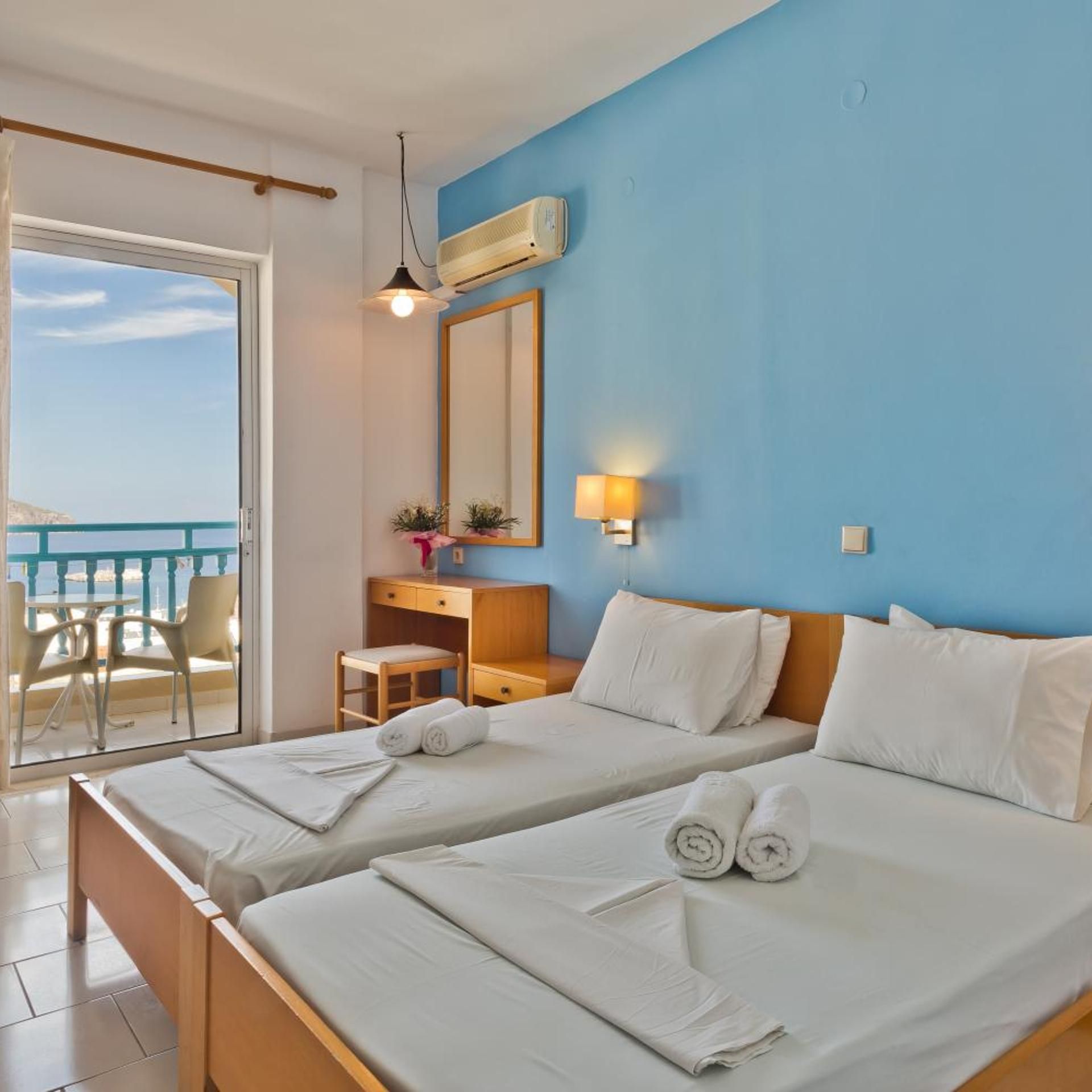 A hotel room with two beds, a desk, a chair, a balcony with a view, and a ceiling fan.