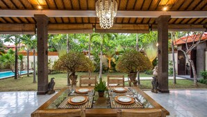 An outdoor dining area with a glass-top table, wooden chairs, and a hanging light fixture. - The Catur Villa (Seminyak)