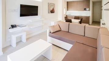 Un salon moderne avec un canapé d’angle, un téléviseur à écran plat, une petite table blanche et un coin cuisine à l’arrière-plan.