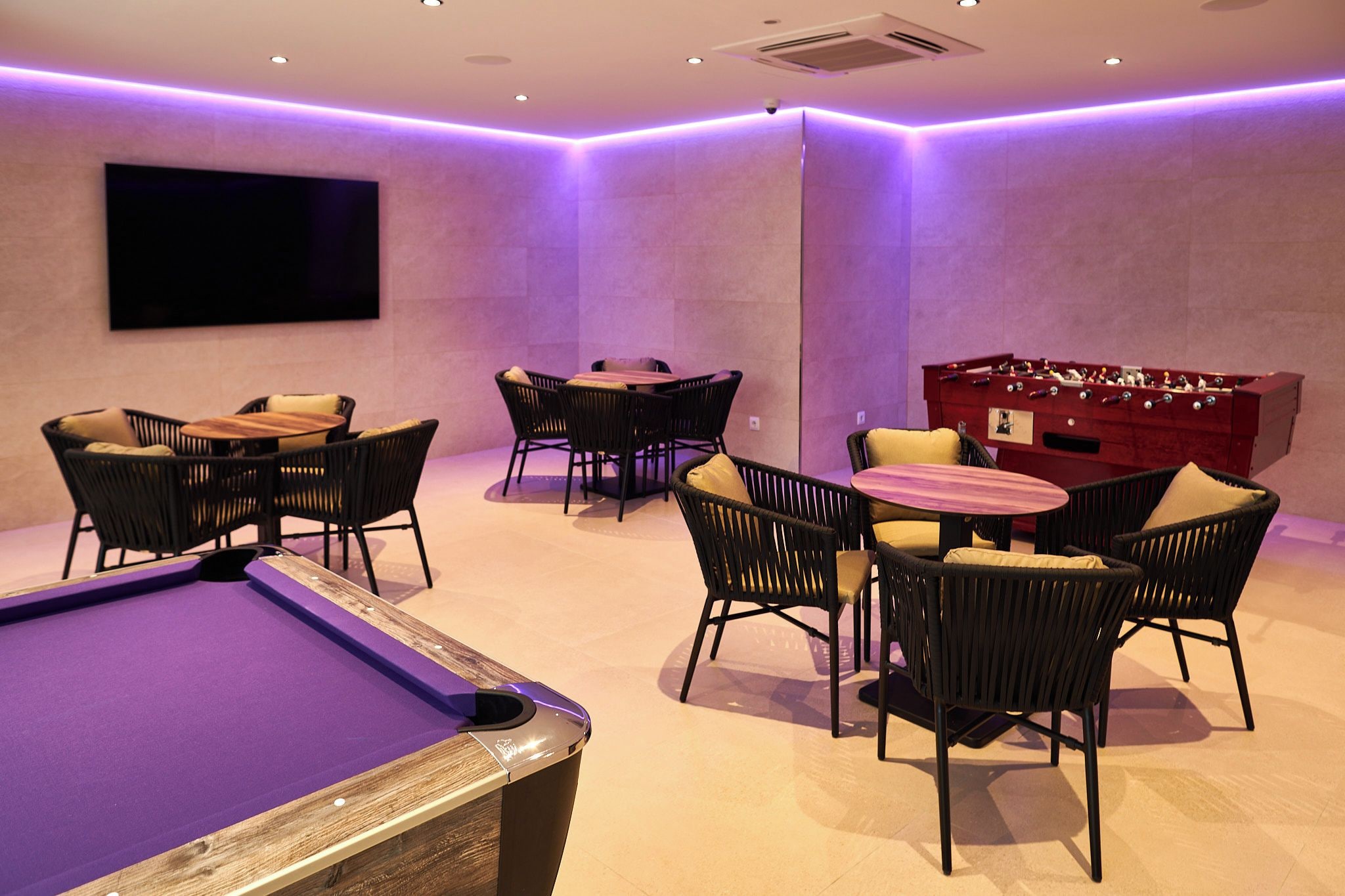 A pool table with surrounding chairs and a foosball table in a room with purple ambient lighting.