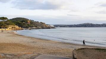 Une plage où une personne se promène le long du rivage, un promontoire rocheux et, au loin, une colline parsemée de bâtiments.