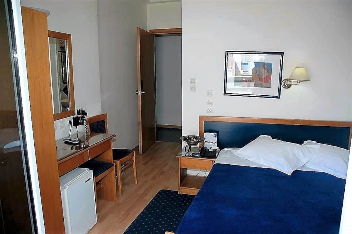 Habitación de hotel con cama, escritorio y silla, un refrigerador pequeño, mesita de noche con lámpara y un cuadro en la pared.