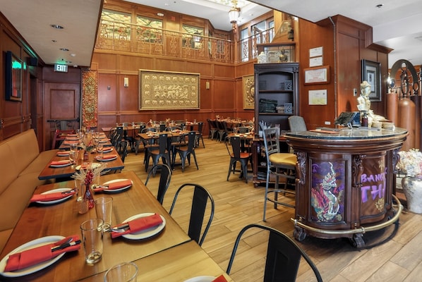 A restaurant with a long wooden table set for dining, a bar area with a decorative counter, and a high ceiling with ornate wood paneling.