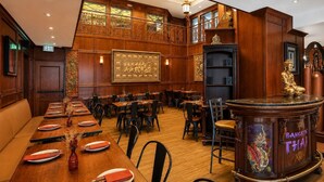 Un restaurante con una larga mesa de madera dispuesta para una comida, una estatua de Buda y un letrero que dice 'Bangkok Thai'.