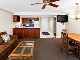 A living room with a sofa, coffee table, and dining table with chairs. A kitchen with wooden cabinets and appliances is visible in the background.