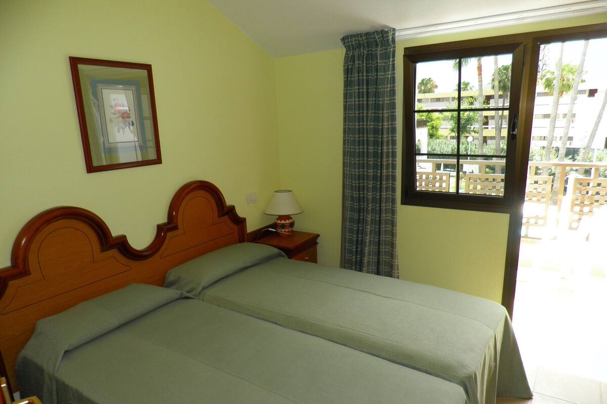 A room with a bed, a nightstand, a lamp, a window with a view of buildings, and a framed picture on the wall.