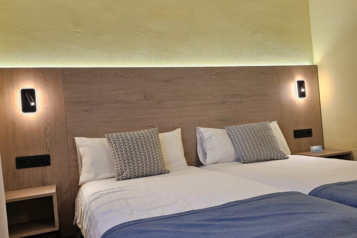 Quarto de hotel com cabeceira de madeira, duas camas com roupa de cama azul e abajures.