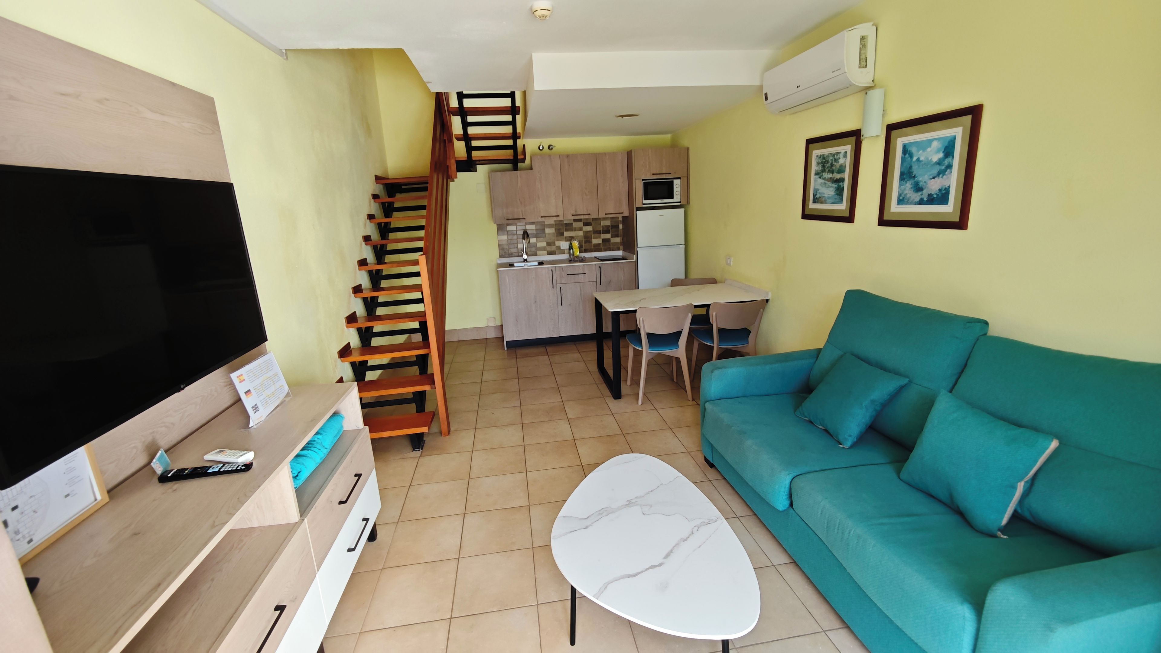 A living room with a TV, sofa, coffee table, and a staircase leading to a kitchen.