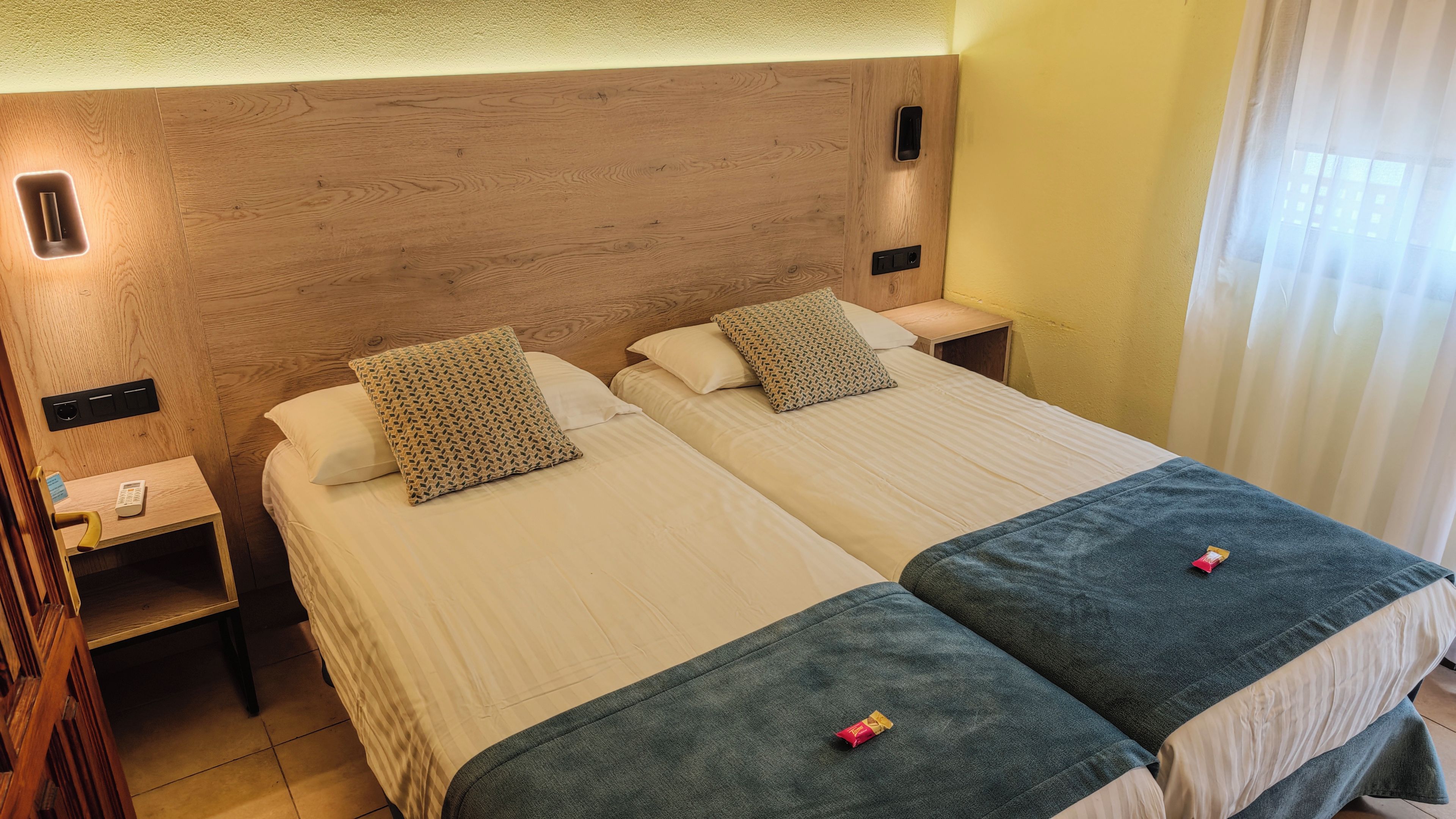 A hotel room with two beds, a wooden headboard, bedside tables with lamps, and a window with curtains.