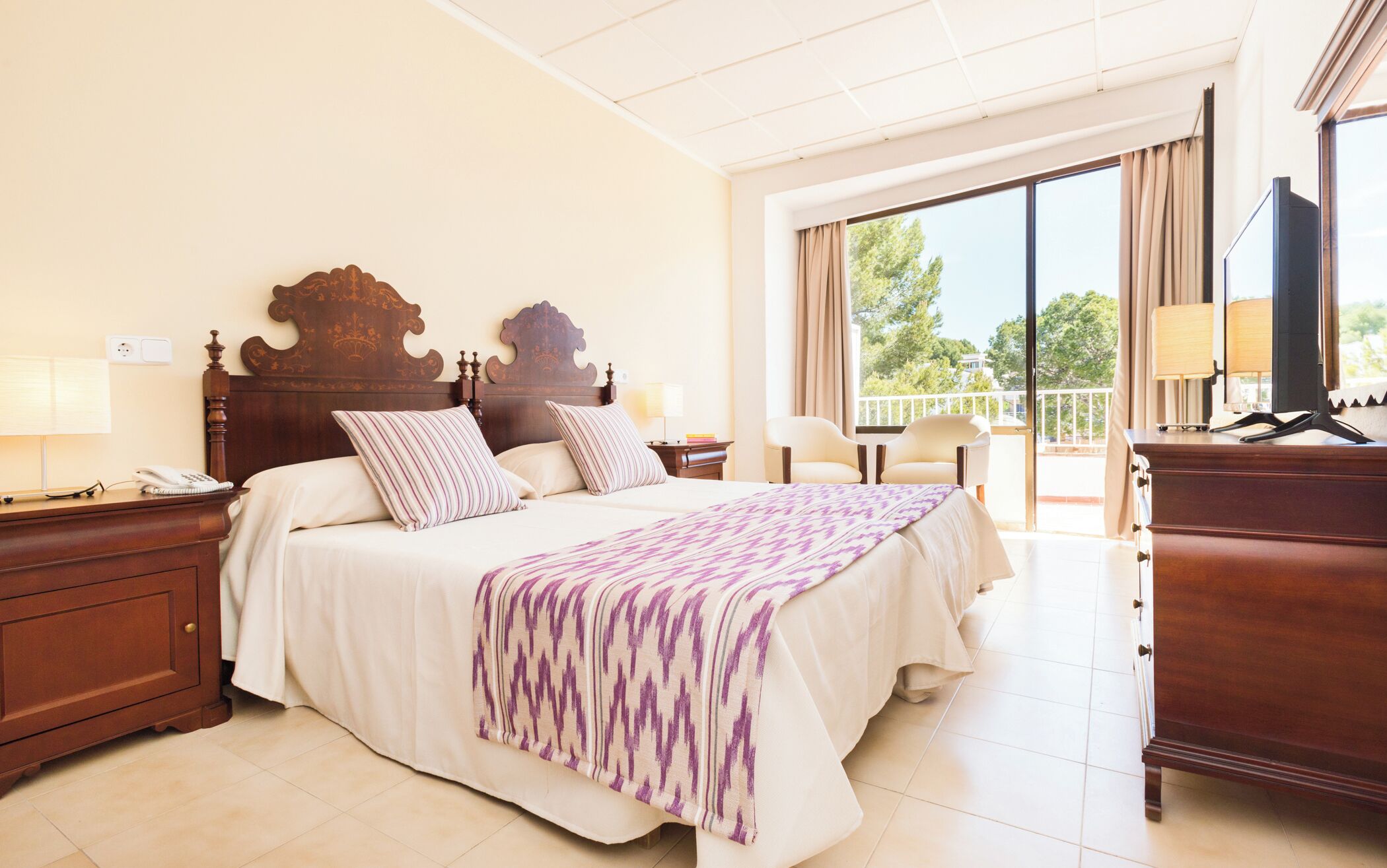 Habitación de hotel con una cama grande, un escritorio con teléfono, una silla, un televisor y un balcón con vistas.