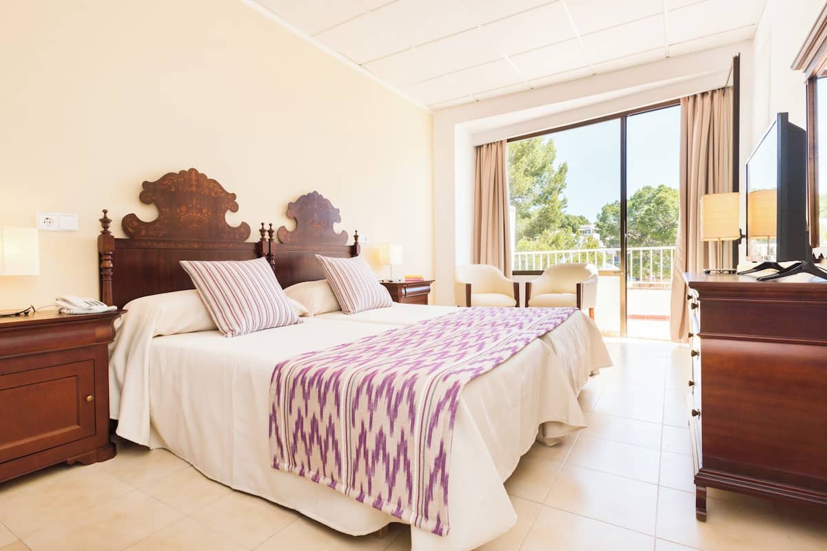 A hotel room with a large bed, a desk with a telephone, a chair, a TV, and a balcony with a view.
