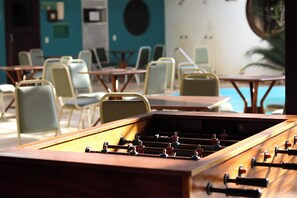 A foosball table in a room with multiple tables and chairs, a pool, and a teal wall. - Guarita Park Hotel By Life Hoteis (Torres)