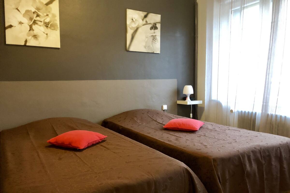 Ein Hotelzimmer mit zwei Betten, jedes mit einem roten Kissen und einer gestreiften Bettdecke. Ein Nachttisch mit einer Lampe steht daneben, und an der Wand hängt ein Kunstwerk.