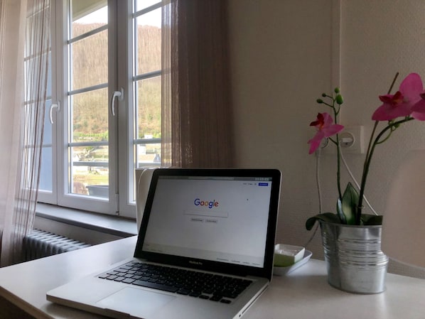 Un MacBook Pro sobre un escritorio con vista a una ventana, un jarrón con flores y un radiador.