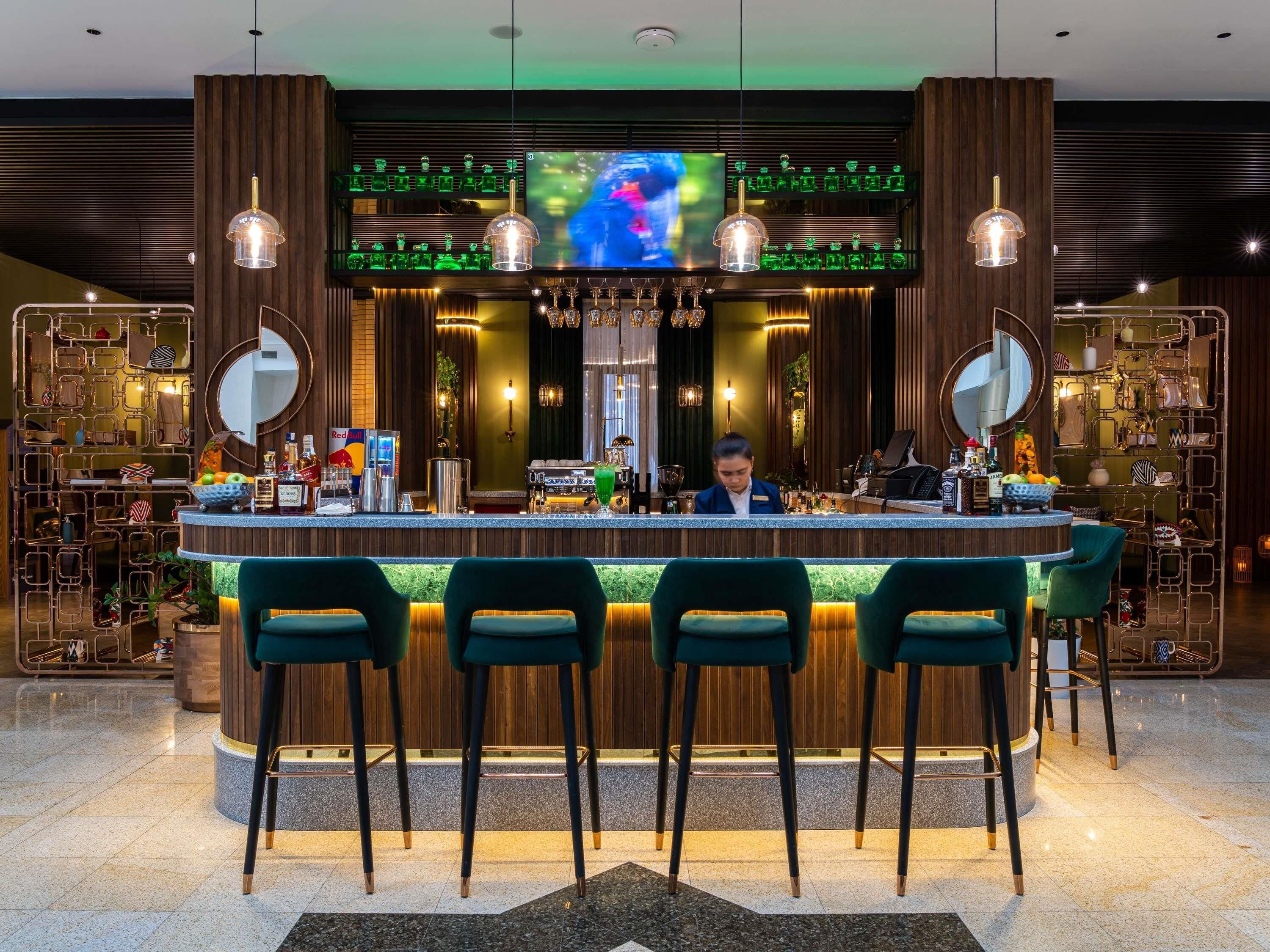 Una zona de bar con un bartender, taburetes verdes, una pantalla de televisión y estantes con botellas y vasos.