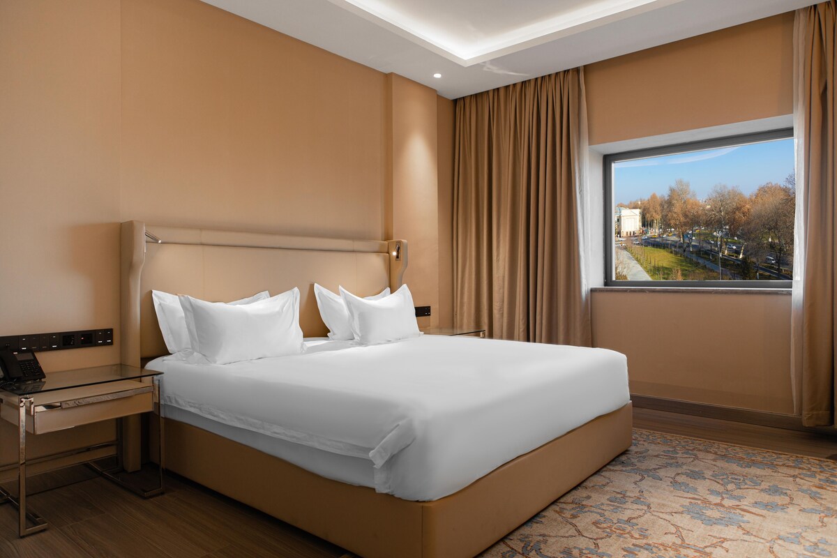 A hotel room with a large bed, a desk with a laptop, and a window with a view of a cityscape.