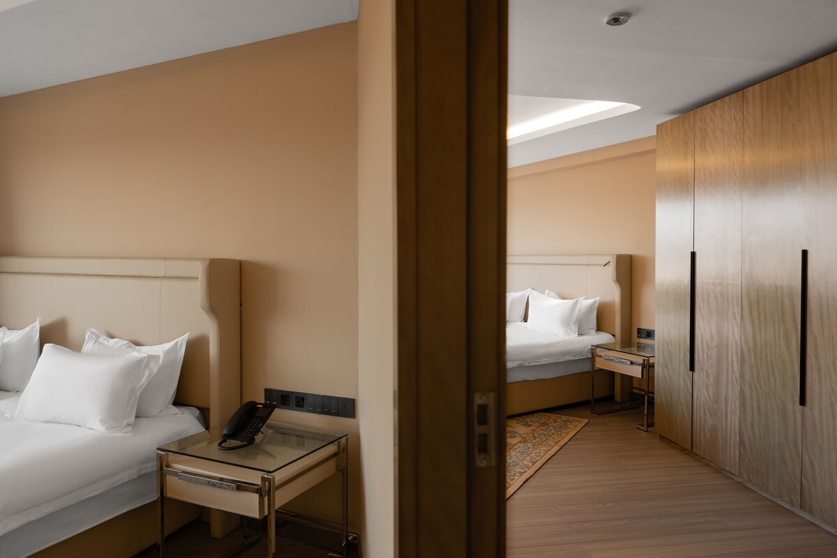 A hotel room with two beds, a bedside table with a phone, a small table with a lamp, and a wardrobe.