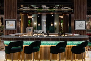 A bar area with a wooden bar counter, green bar stools, and framed artwork on the walls. - Mövenpick Samarkand (Samarkand)