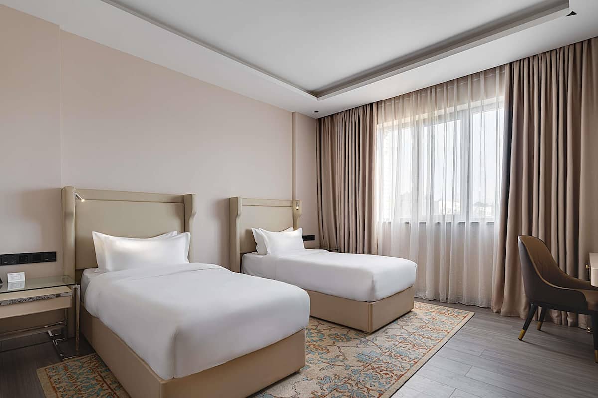 A hotel room with two beds, a desk, a chair, and a large window with curtains.