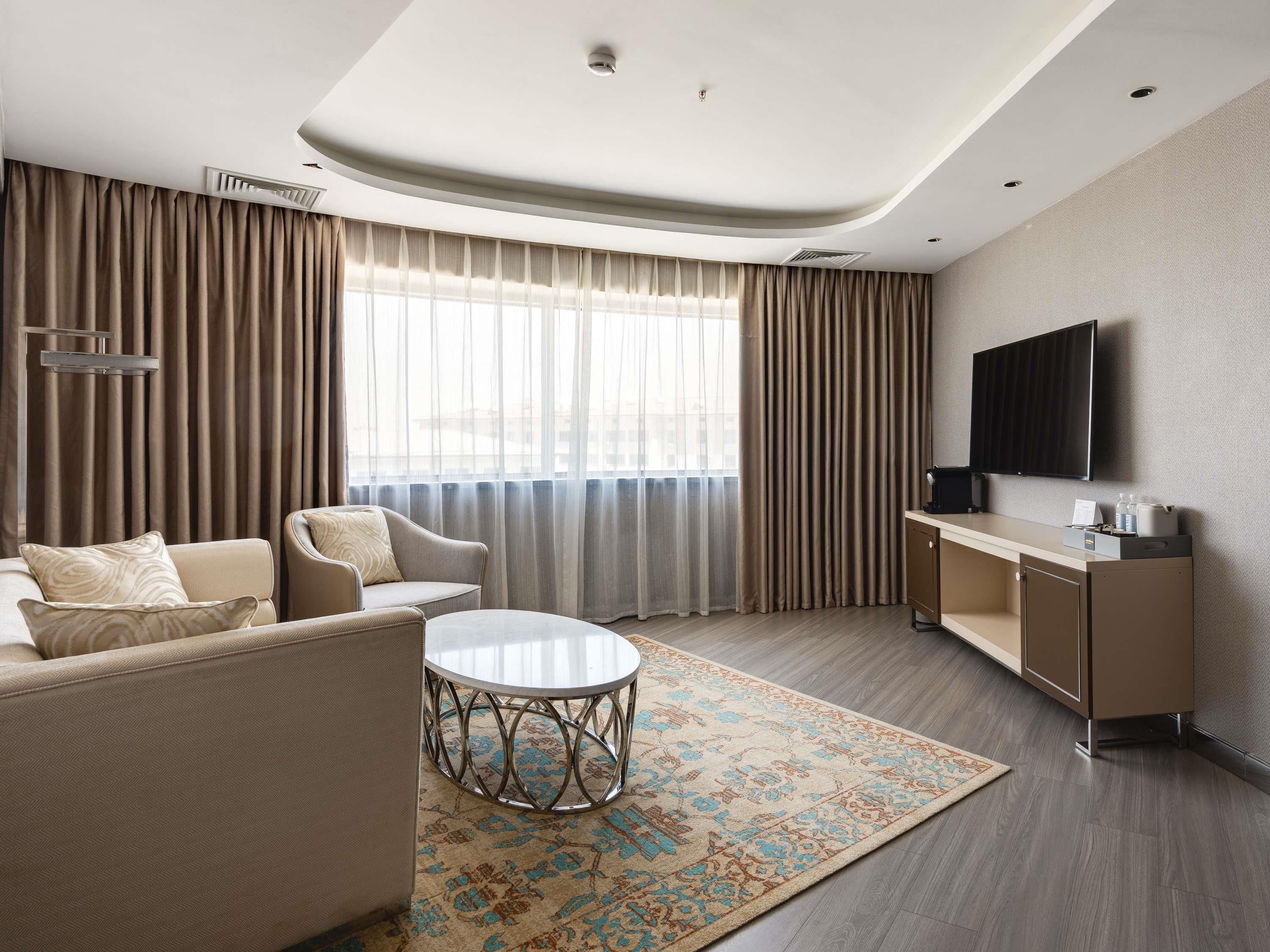 Una habitación de hotel moderna con sofá, sillón, una mesita redonda y un televisor de pantalla plana montado en la pared.