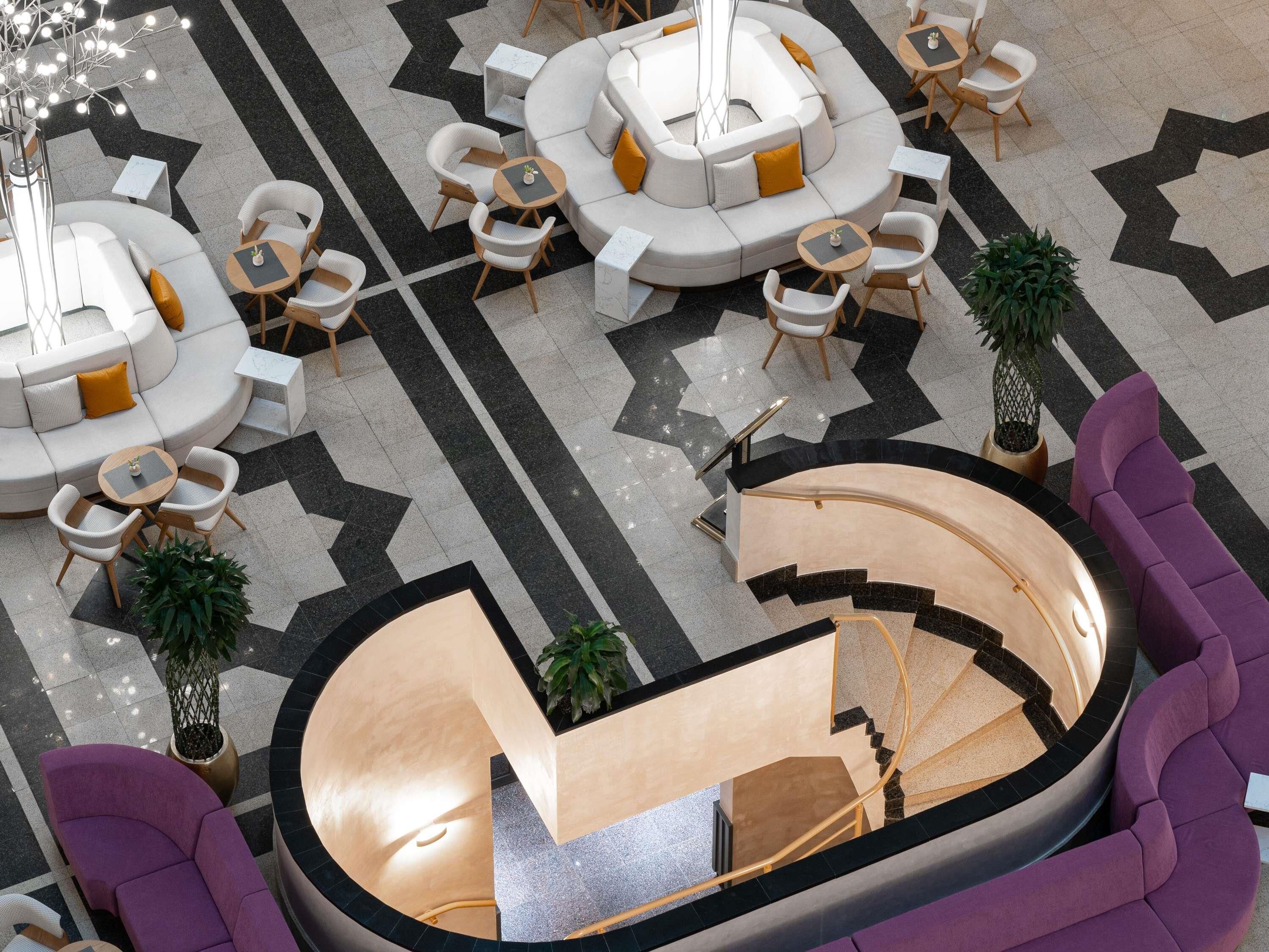 A modern hotel lobby with a central seating area, multiple tables, and a staircase leading to an upper level.