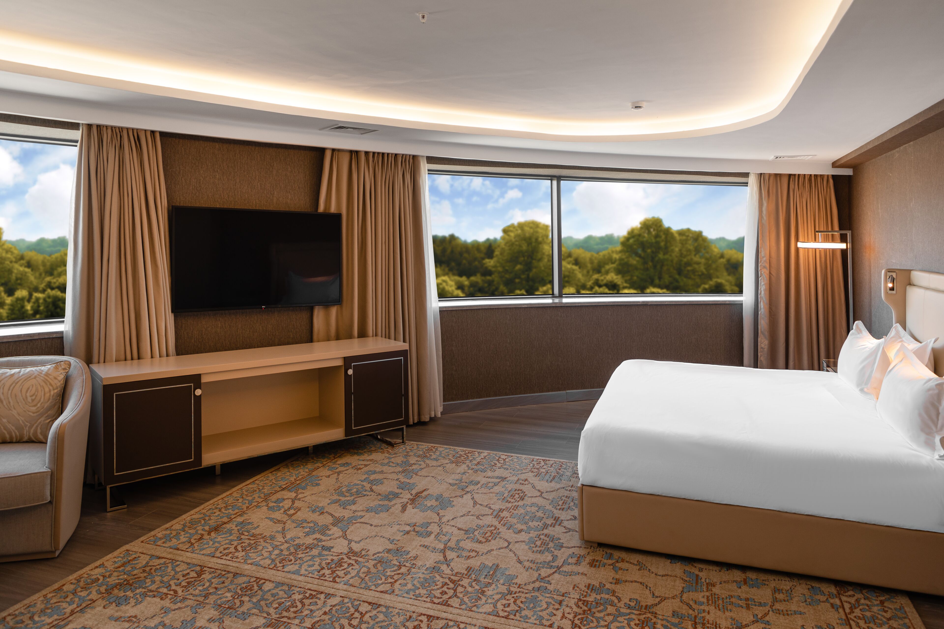 A modern hotel room with a large bed, a flat-screen TV mounted on the wall, a seating area with a sofa and a desk, and a large window offering a scenic view.