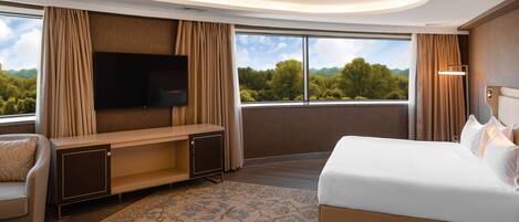 A modern hotel room with a large bed, a flat-screen TV mounted on the wall, a seating area with a sofa and a desk, and a large window offering a scenic view.
