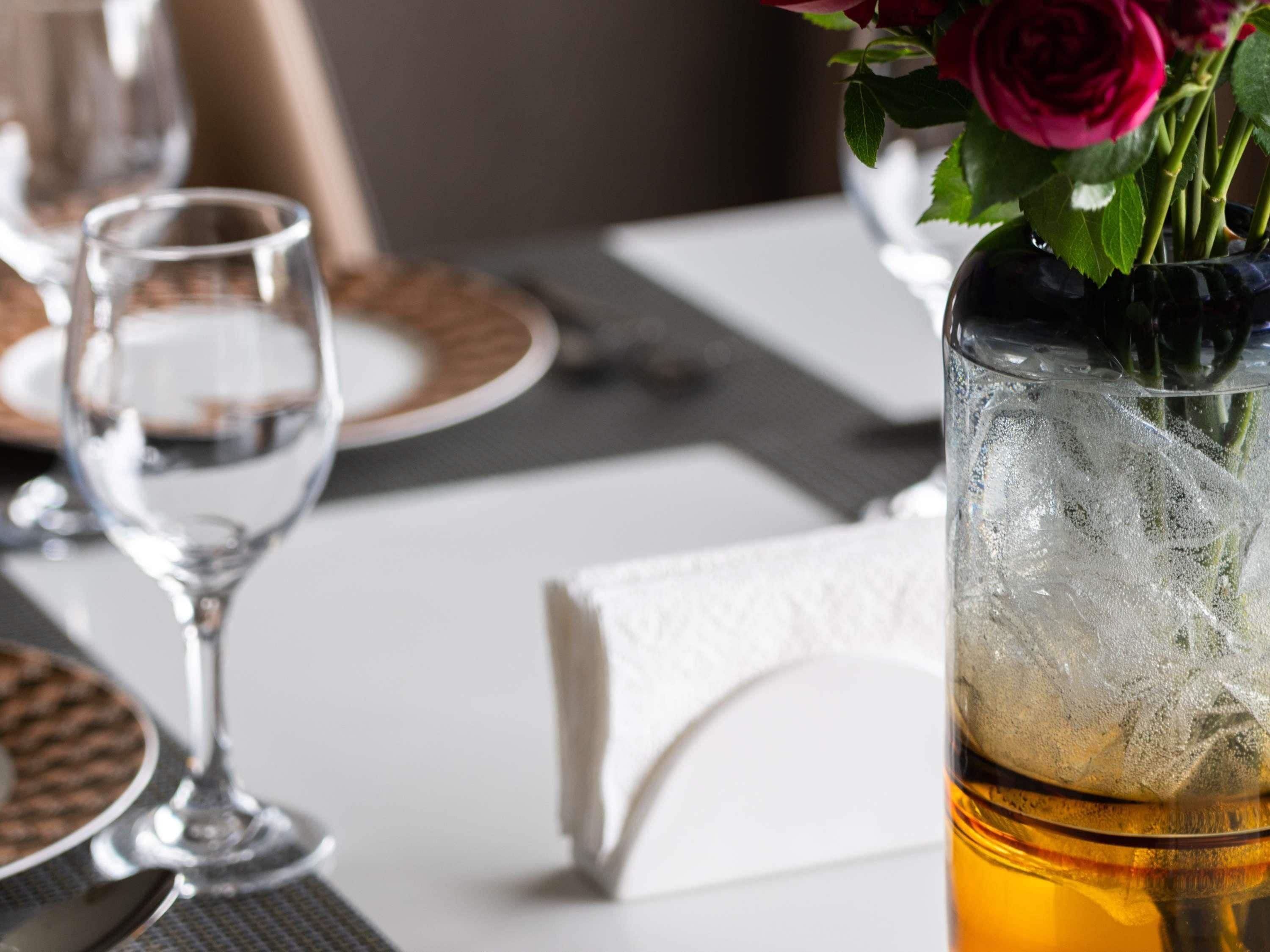 Una mesa de comedor con un vaso de líquido ámbar, cubitos de hielo y un ramillete de rosas rojas.