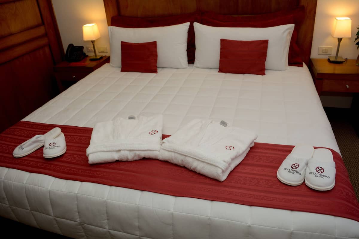 A neatly made hotel bed with red and white bedding, a red throw blanket, and a set of white slippers and towels with a logo.
