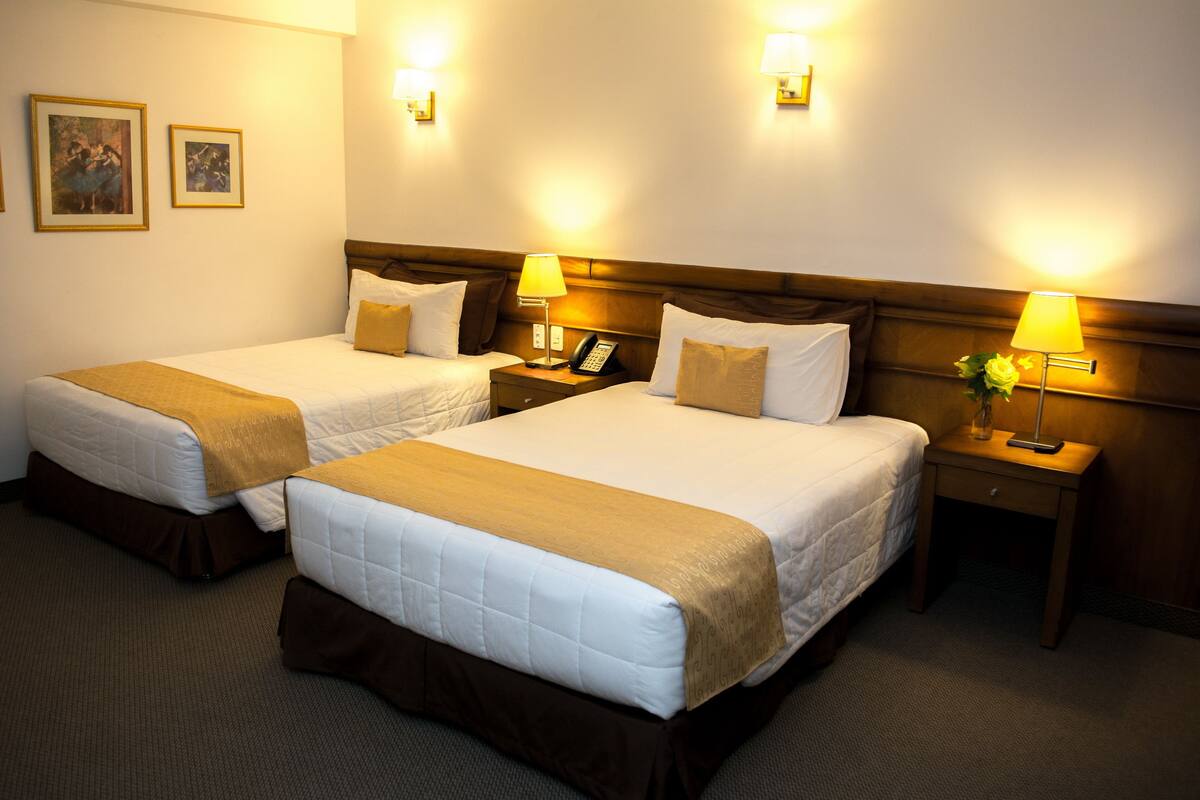A hotel room with two beds, each with a brown headboard, white bedding, and a brown throw blanket. There are bedside lamps, a telephone, and a small table with a vase of flowers.