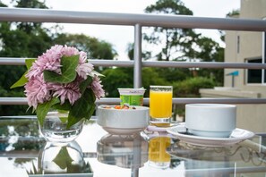 A breakfast setup on a balcony with a glass table, a bowl of cereal, a glass of orange juice, a cup, and a vase of flowers.
