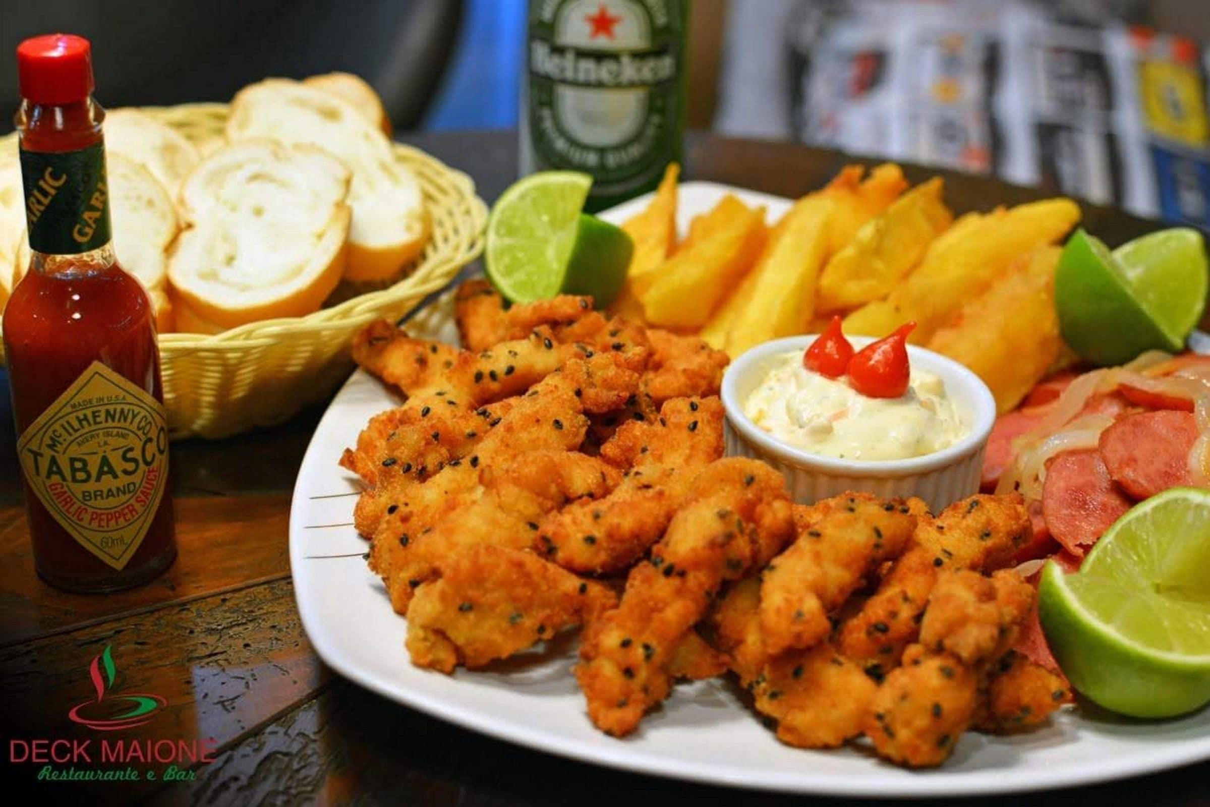 Une assiette de poisson frit, de frites et de saucisses en tranches, accompagnée d’une sauce tartare et de quartiers de citron vert.