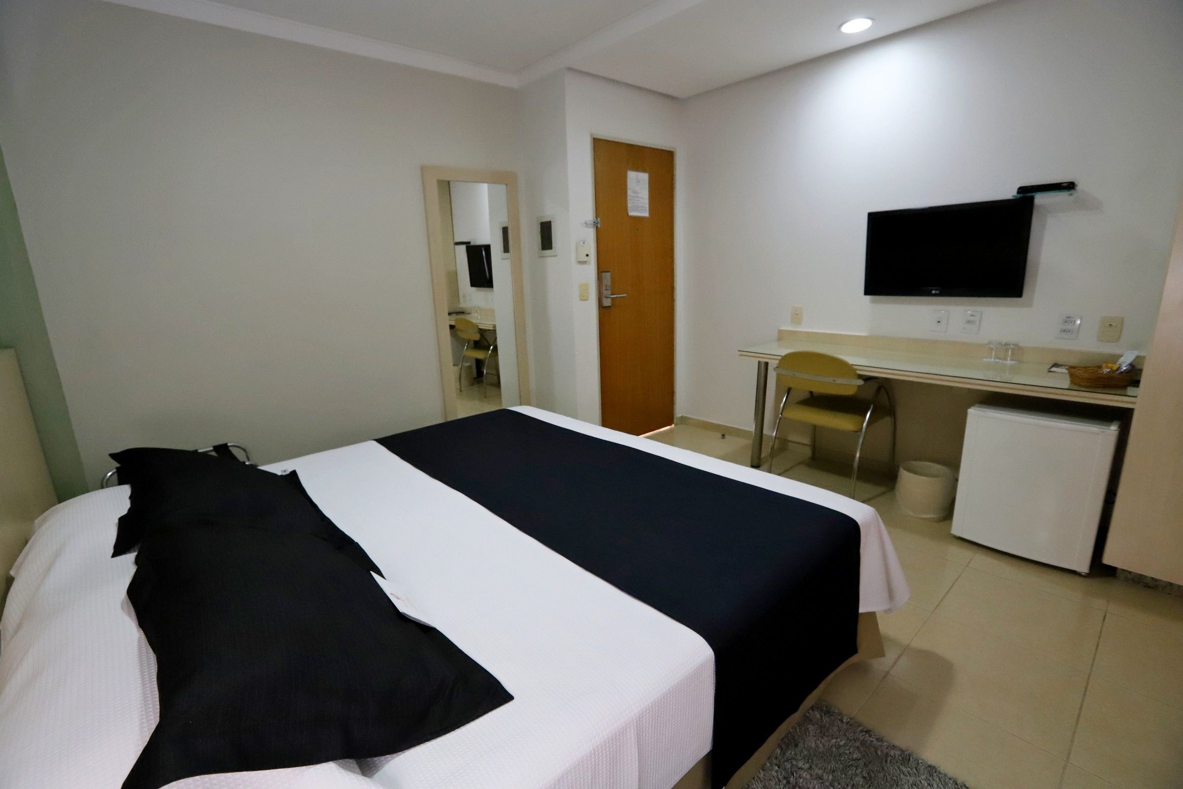 Quarto de hotel com cama, escrivaninha, cadeira, televisão e pequeno frigorífico.