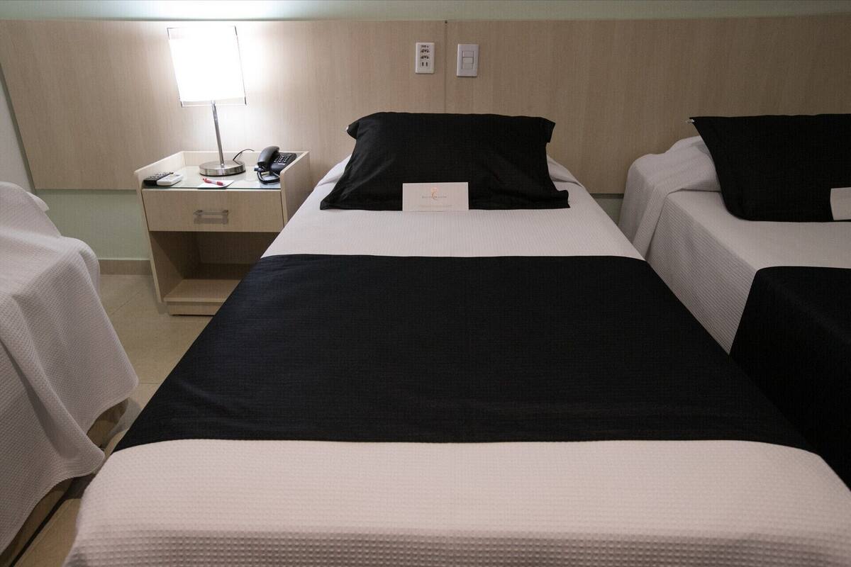 A hotel room with three beds, each with a black and white bedspread. There is a bedside table with a lamp and a phone.