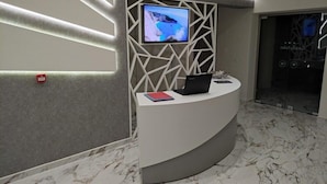 A reception desk with a computer, a TV mounted on a geometric wall, and a marble floor. - Eleni's Garden & Castello Suites (Zakynthos)