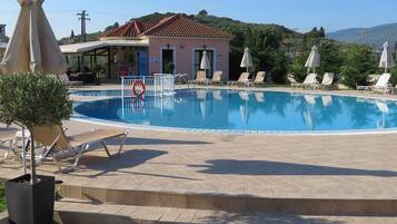 A swimming pool with lounge chairs and umbrellas, a building with a tiled roof, and a hilly landscape in the background.