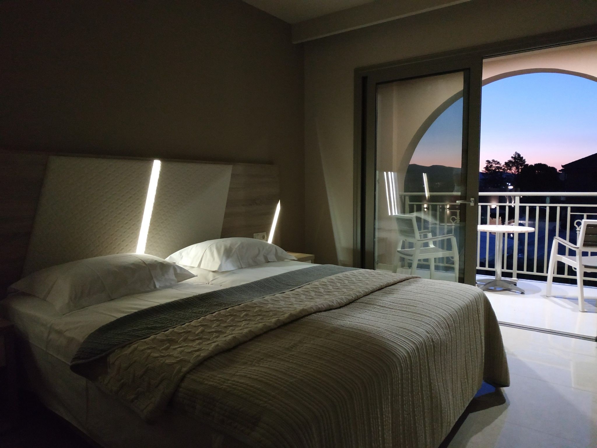 A hotel room with a bed, a large window, and a balcony with chairs.
