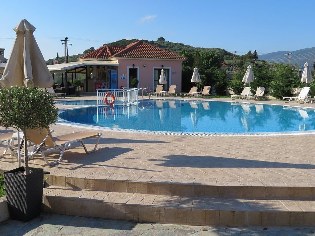 A swimming pool with lounge chairs and umbrellas, a building with a tiled roof, and a hilly landscape in the background.