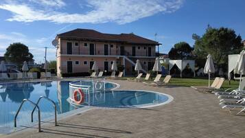 A swimming pool with lounge chairs and umbrellas, a lifebuoy, and a building with balconies in the background.