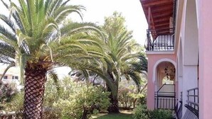 A building with a pink facade, a balcony with black railings, and a garden with palm trees and shrubs. - Eleni's Garden & Castello Suites (Zakynthos)
