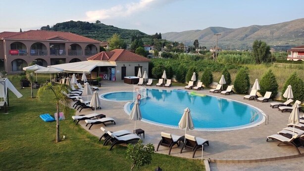 A pool area with lounge chairs and umbrellas, a building with a red roof, and a hilly landscape in the background. - Eleni's Garden & Castello Suites (Zakynthos)