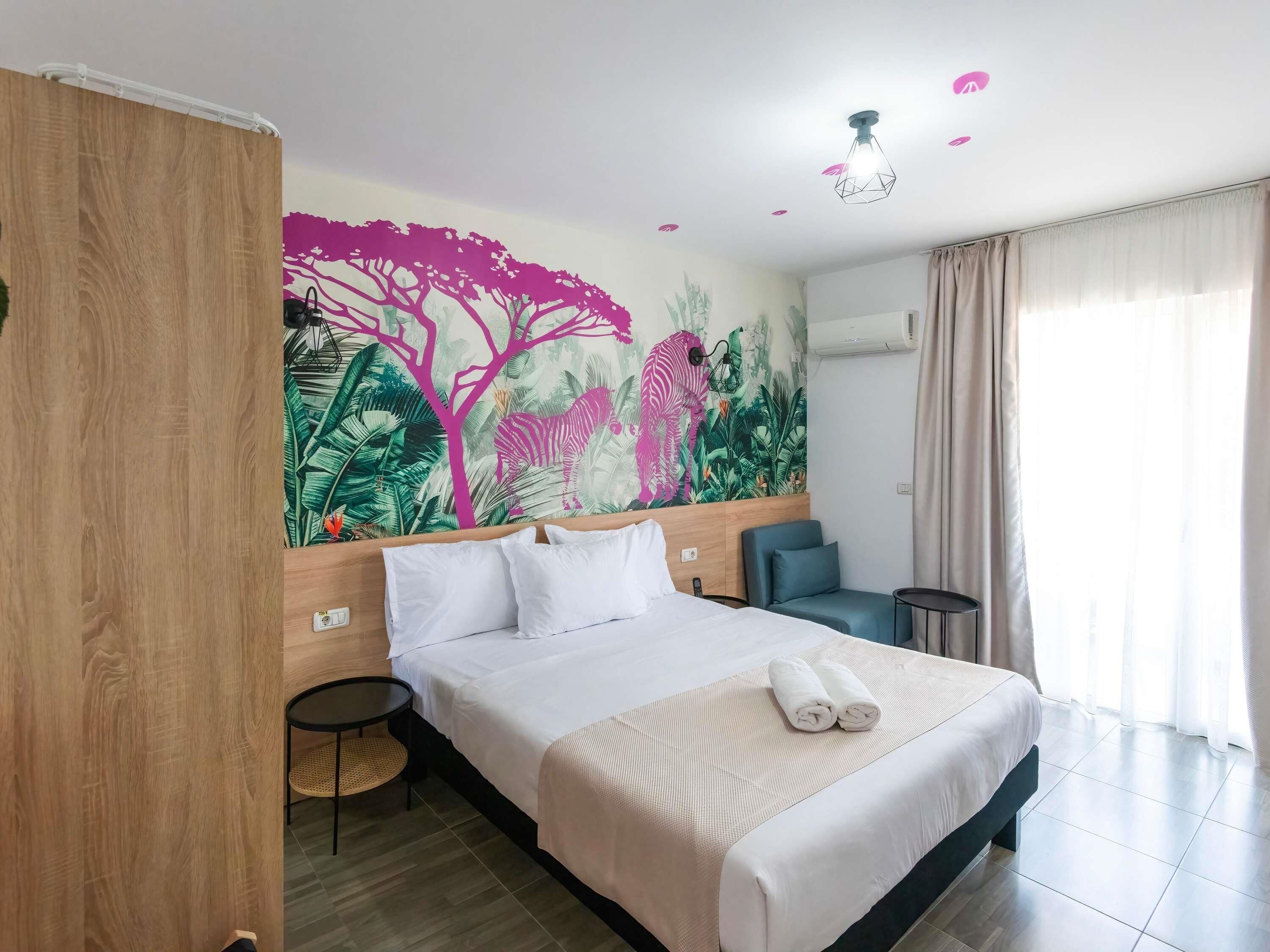A hotel room with a large bed, a small desk, a chair, a wall mural, and a window with curtains.
