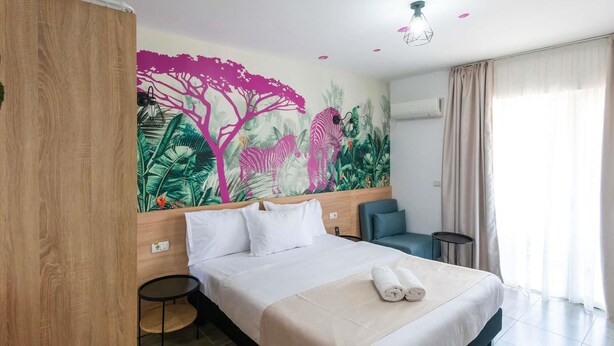 Habitación de hotel con una cama grande, un escritorio pequeño, una silla, un mural en la pared y una ventana con cortinas.