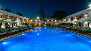 A swimming pool with lounge chairs and umbrellas, surrounded by buildings with arched windows and warm lighting. - ibis Styles Venus (Opening June 2024) (Venus)