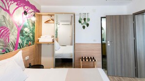 A hotel room with a bed, a wardrobe, a small wooden stool, a hanging plant, and a wall with a tropical mural. - ibis Styles Venus (Opening June 2024) (Venus)