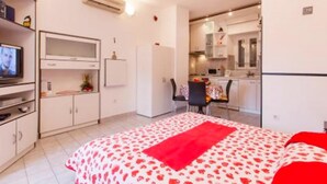 A compact bedroom with a bed, a television, a desk, and a kitchenette.